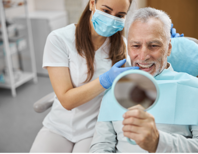 The professional is in a white uniform and blue gloves, while the patient, seated in a dental chair with a blue bib, holds up a mirror. The background shows dental equipment and shelves, emphasizing the clinical environment.