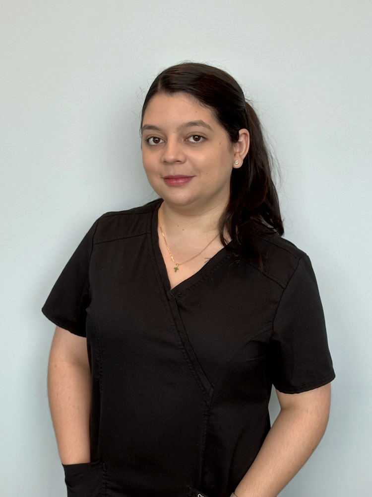 The image shows a woman wearing a black scrub top and a necklace with her hands on the pocket, against a plain light-colored background.