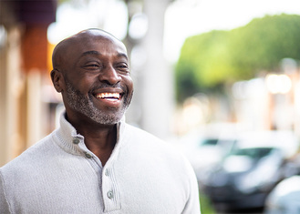 A smiling middle-aged man with a bald head and grey beard, wearing a light grey sweater. He is outdoors with a blurred background of trees and cars, enjoying a bright day.