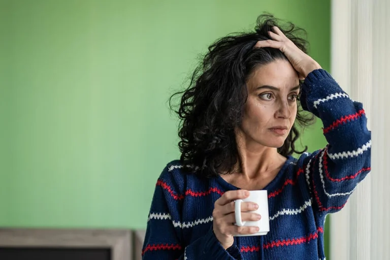 A woman with dark curly hair wearing a blue striped sweater holds a white mug and rests one hand on their head. She is experiencing dental anxiety